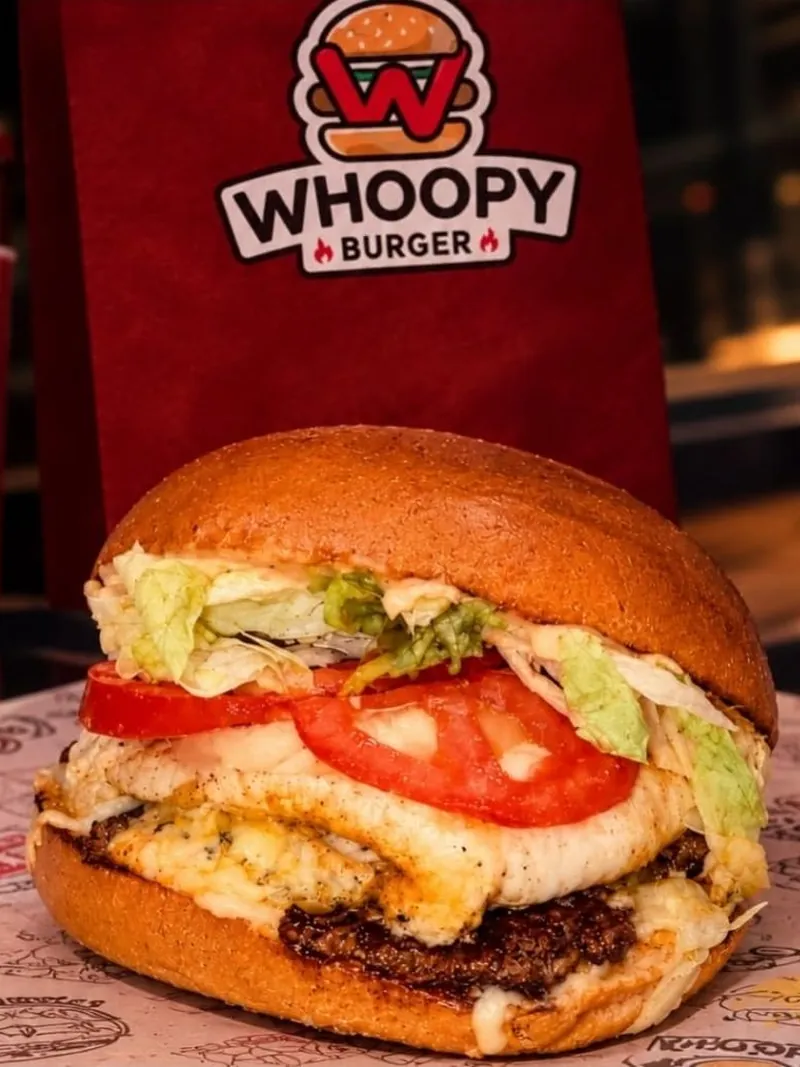 Un burger au pain brioché garni de salade, de tranches de tomate, de fromage fondu et d’un steak haché, posé sur un papier imprimé devant un sac rouge Whoopy Burger