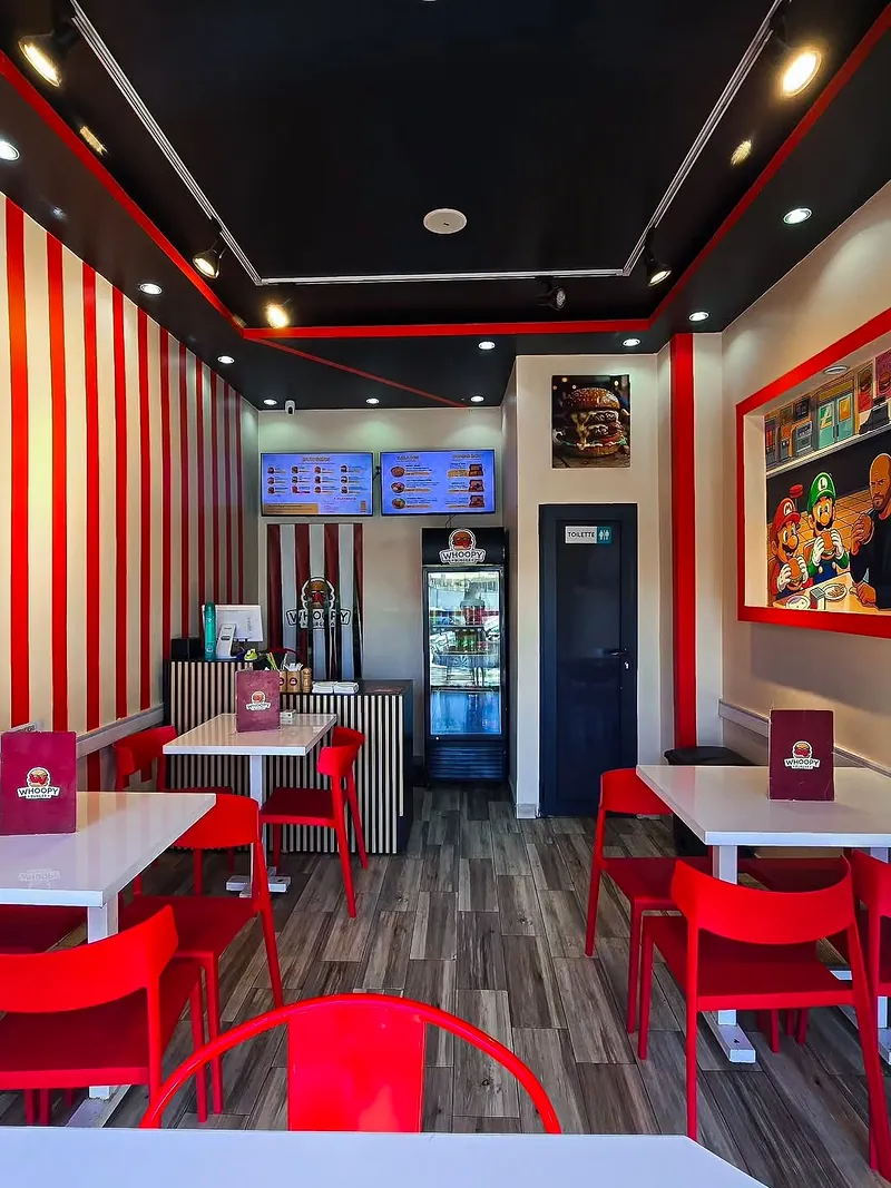 La salle de Whoopy Burger : plafond sombre, rayures rouges et blanches, tables blanches et chaises rouges